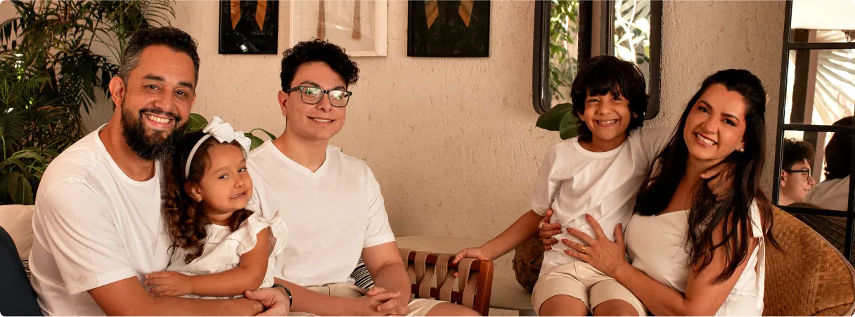 Família de quatro pessoas, pai, mãe, dois filhos e uma filha sentados em um sofá em ambiente interno, todos sorrindo e vestindo roupas brancas.