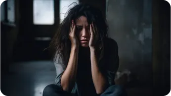 Mulher sentada no chão em um ambiente escuro, com as mãos na cabeça em sinal de desespero.
